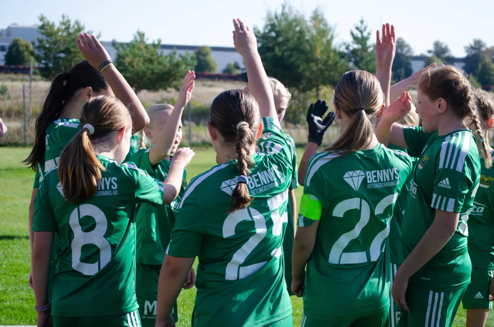 Ungdomslag i gröna matchtröjor sträcker upp händerna i en lagsamling på fotbollsplanen
